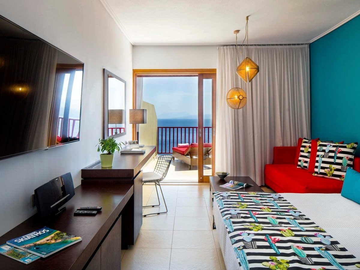 A long room with a bed, a red sofa against a turquoise wall, a run of dark desks and a balcony with a sunbed beyond the doors.