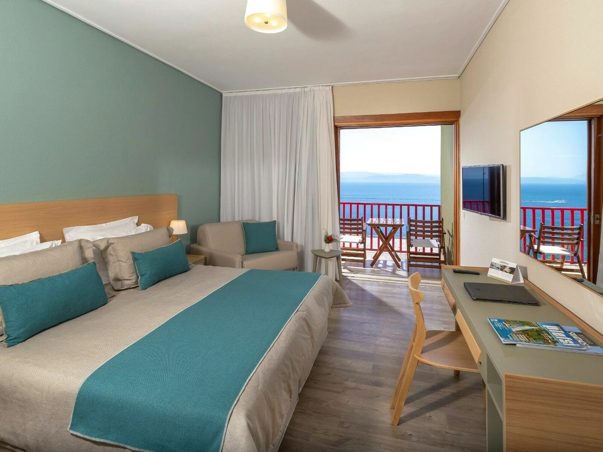 A double room with a green feature wall, a teal bed runner, a small sofa and a desk, opening onto a balcony with wooden chairs above the sea.