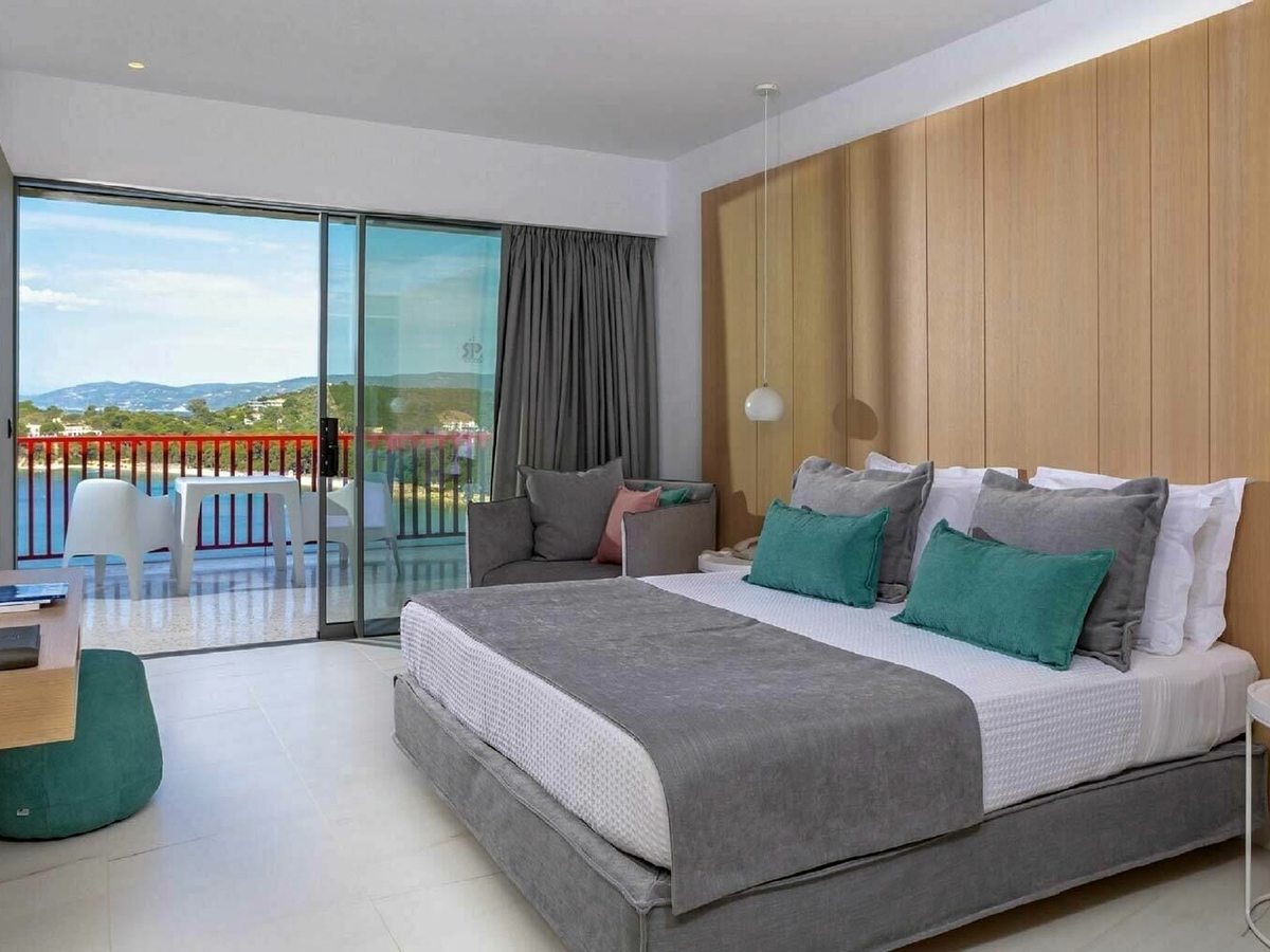 A double room with a pale wood headboard wall and grey and teal bedding, with full-height glass doors to a veranda set with a table and chairs above the bay.