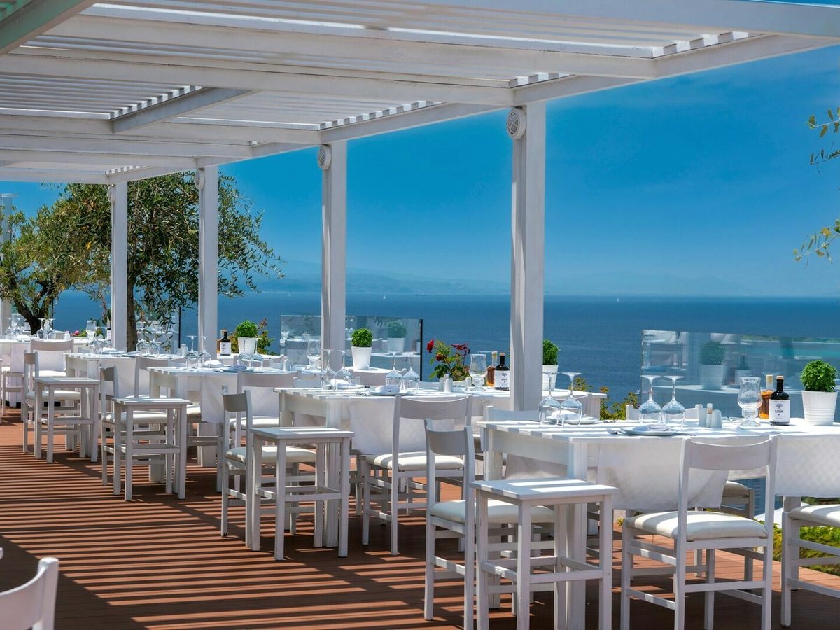 A restaurant terrace under a white slatted pergola, with white tables and chairs set with glasses on timber decking and open sea beyond.