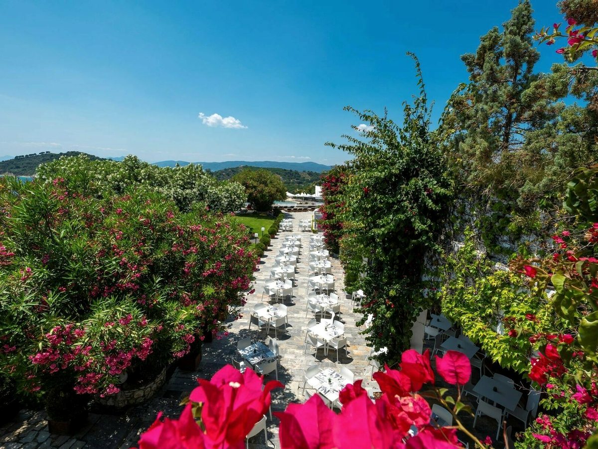 A stone-paved garden walk lined with white tables and chairs, framed by flowering oleander and bougainvillea, with wooded hills and the sea beyond.