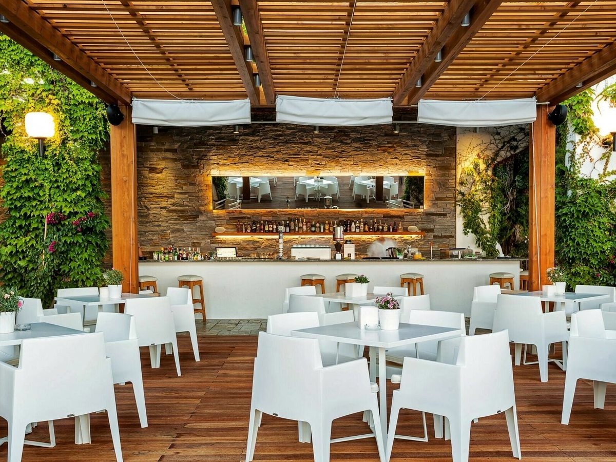 An outdoor bar under a timber pergola, with a stone-faced counter and wooden stools, white tables and chairs, and climbing greenery on either side.