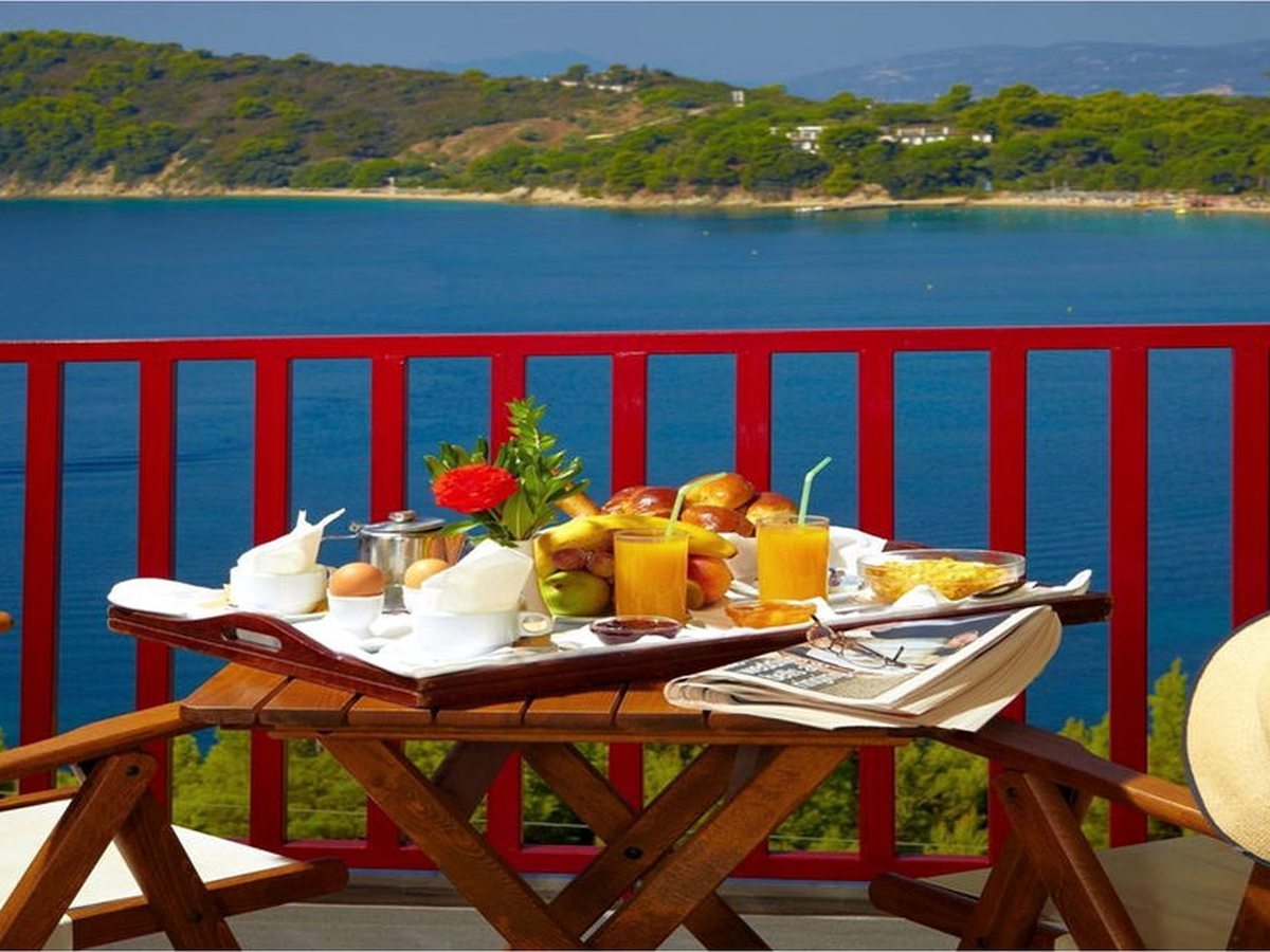 A breakfast tray of bread, fruit, boiled eggs and orange juice on a wooden balcony table, with a red railing and the blue bay behind.