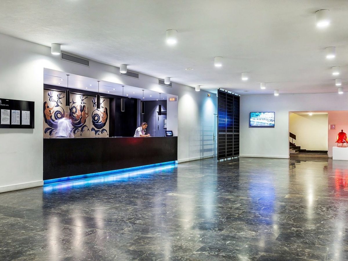 A wide lobby with a grey marble floor and a long dark reception desk lit from below in blue, a receptionist behind it and a painted mural on the wall.