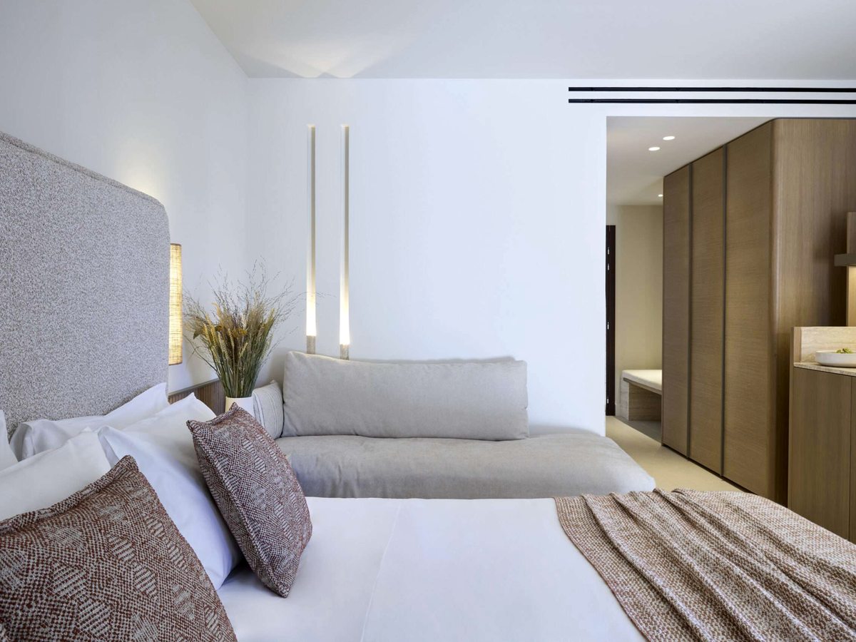 A double bed with a pale grey upholstered headboard, white bedding and a woven rust throw, a long grey sofa along the wall and a fitted oak wardrobe by the entrance.
