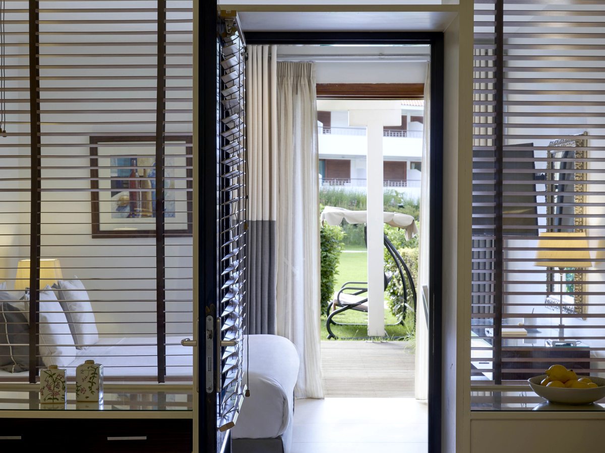 An open door and slatted timber screen inside a room, looking out to a lawn with a cushioned swing seat and a shaded daybed, and a white balconied building beyond.