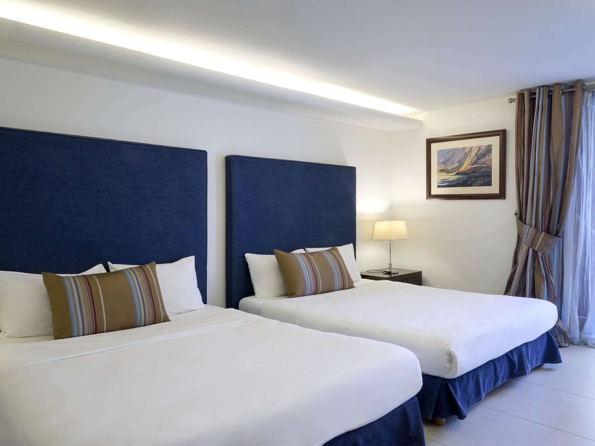 Two double beds side by side under tall navy padded headboards, with striped bolster cushions, a bedside lamp between them and a framed painting on the wall.