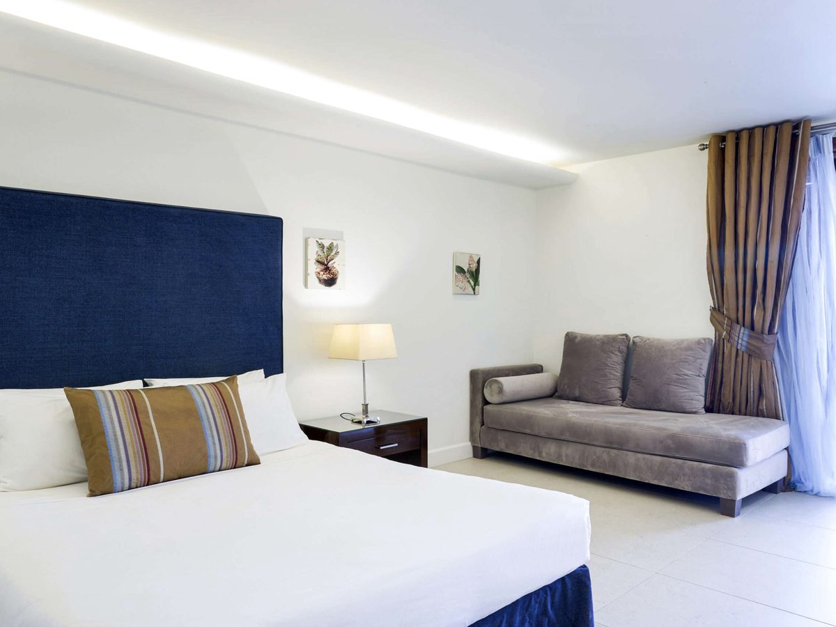 A double bed with a tall navy padded headboard and a striped bolster cushion, a grey velvet daybed sofa against the far wall and long curtains at the window.