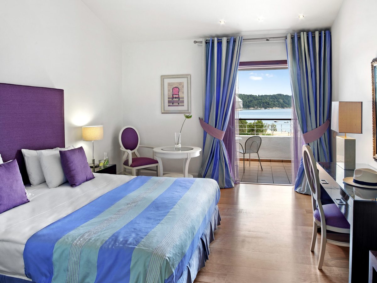 A double bed with a purple padded headboard and a blue striped throw, a small round table and chair, and open balcony doors looking out to a wooded headland and the bay.