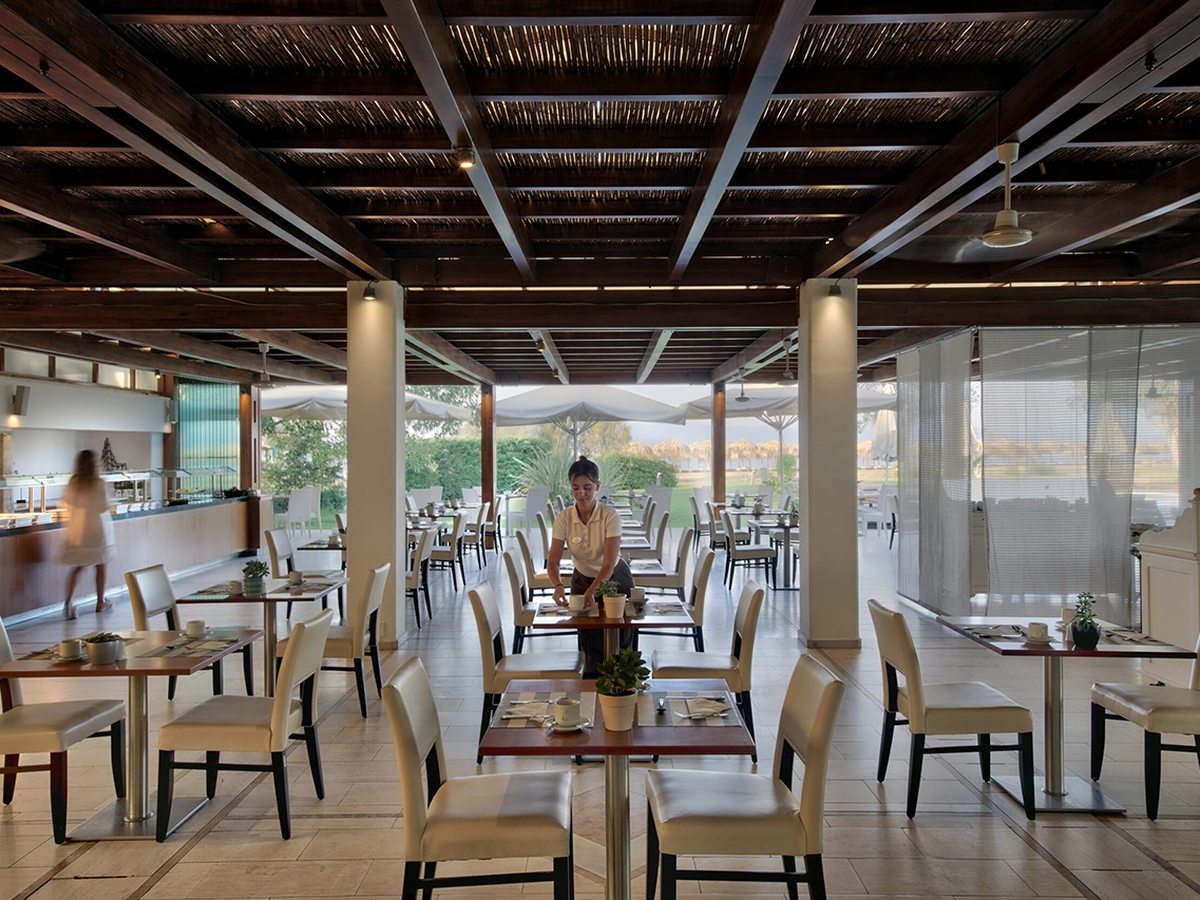 An open-sided restaurant under a dark timber and reed ceiling, with set tables and cream chairs, a buffet counter along the left wall and a lawn and beach parasols outside.