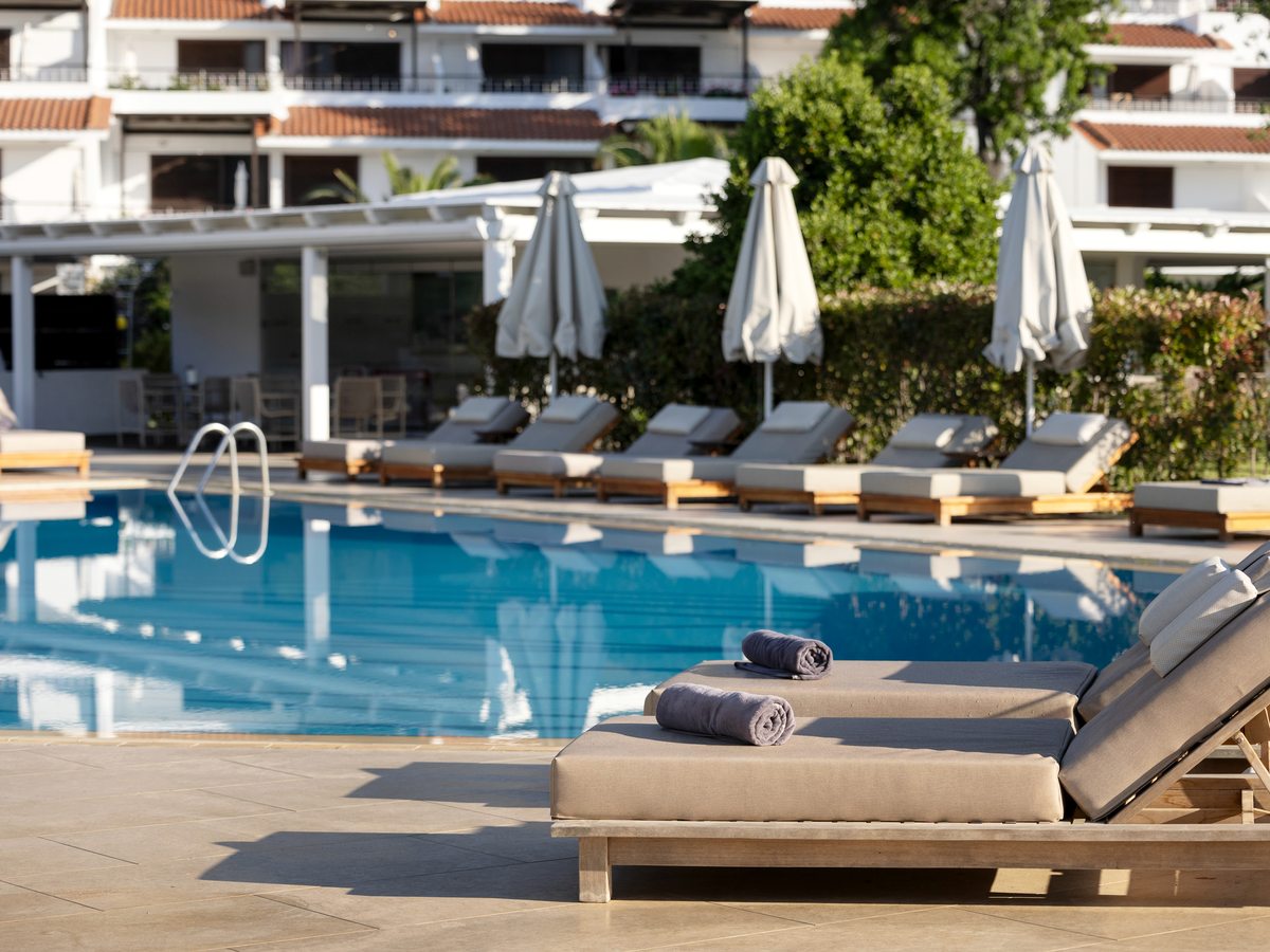 A rectangular pool with wooden-framed sun loungers, rolled towels and furled white parasols along the paved surround, and white guest-room buildings with red-tiled roofs behind.