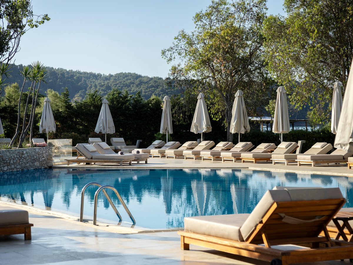 A large pool with a long row of wooden sun loungers and furled cream parasols along the far side, backed by trees and a pine-covered hillside.