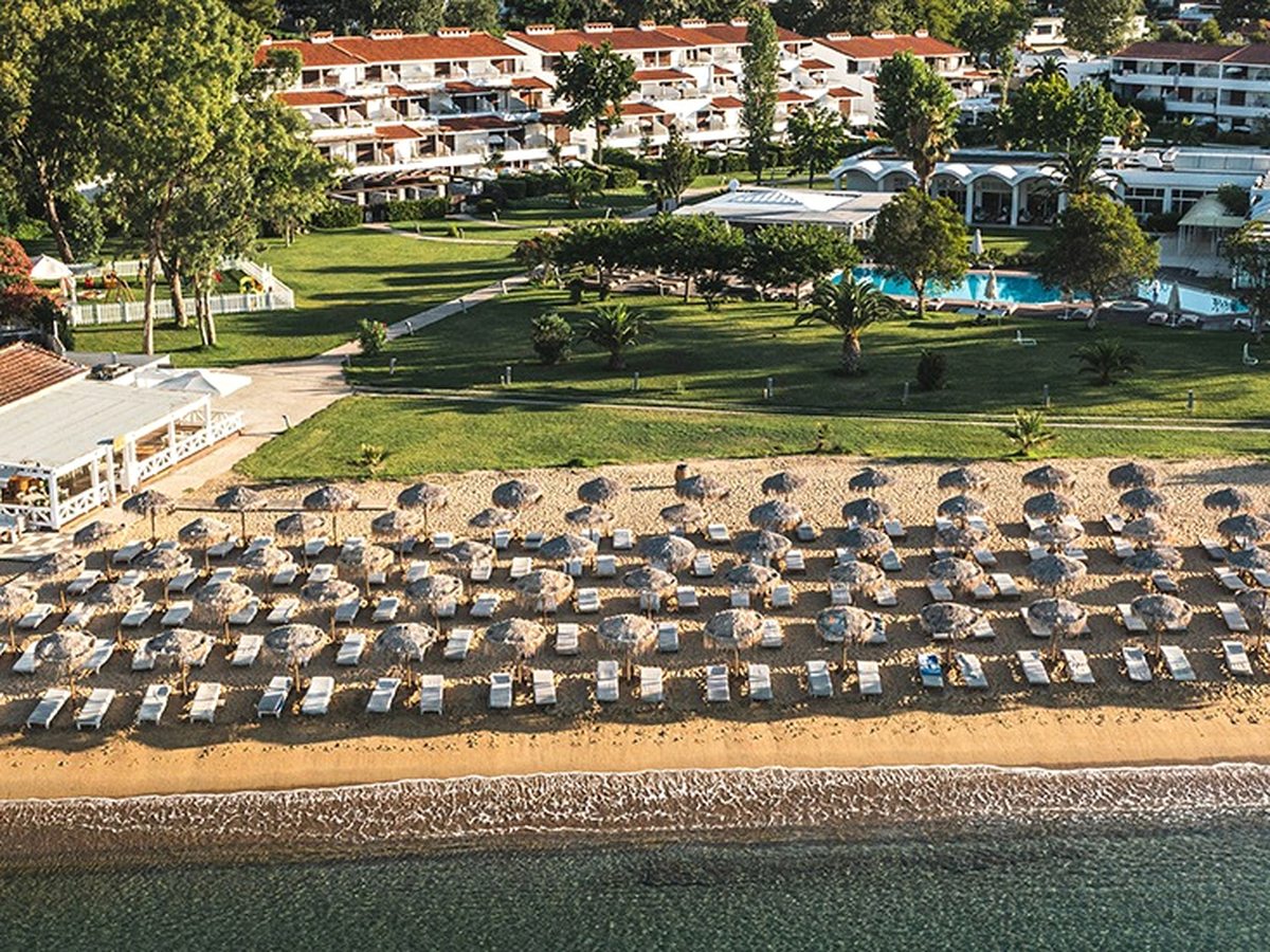 Aerial view of low white buildings and lawns behind a long sandy beach packed with rows of thatched umbrellas and sun loungers at the water's edge.
