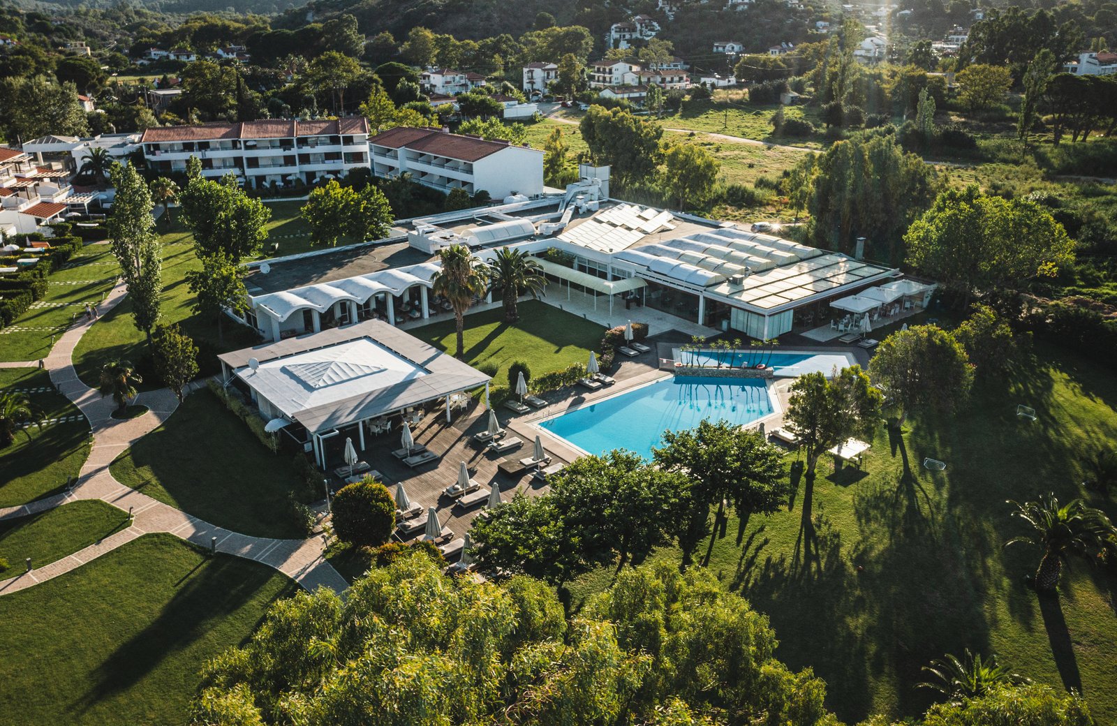 Aerial view of Skiathos Princess Resort: white buildings and two blue pools set among lawns, paths and trees, with village houses and wooded hills rising behind.