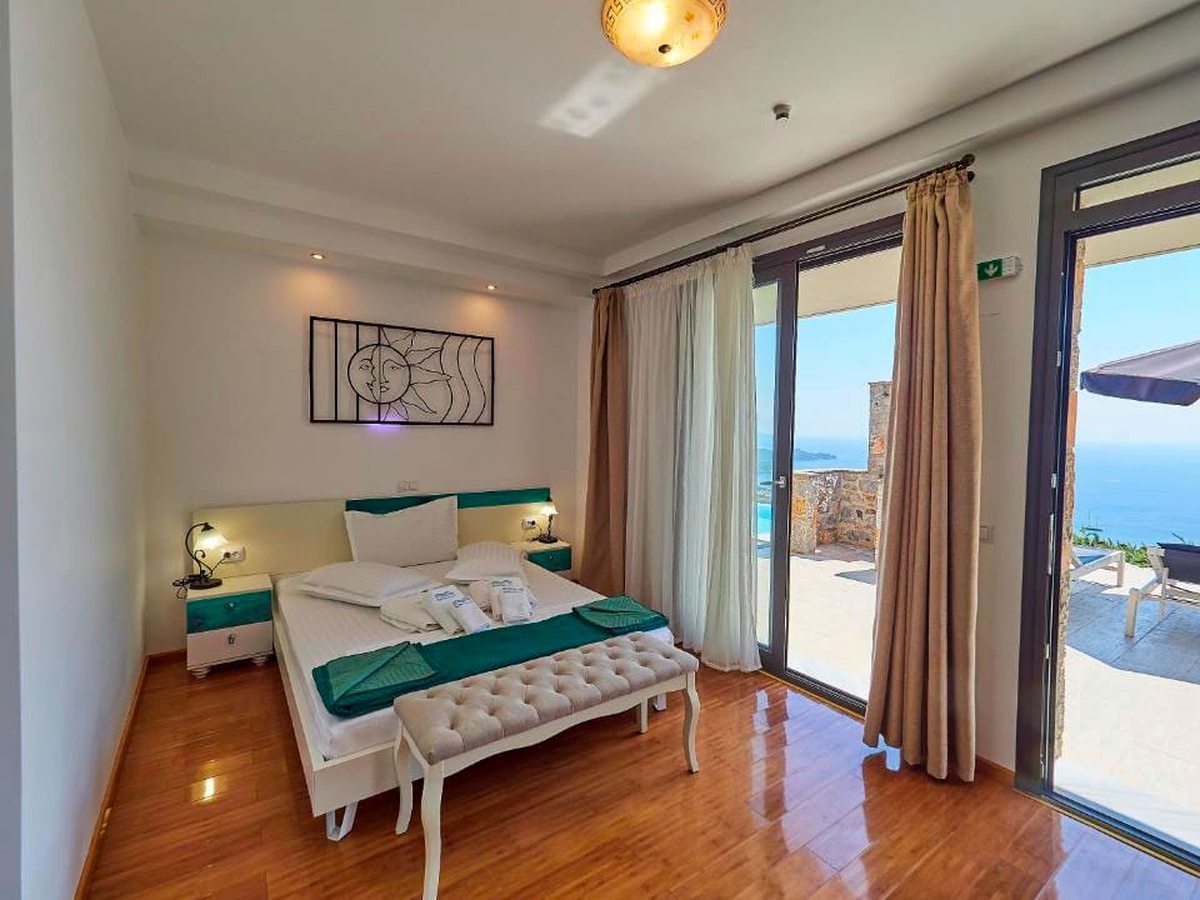 A bedroom with a white double bed, teal bedside tables and a polished wood floor, with sliding glass doors onto a terrace and the sea beyond.