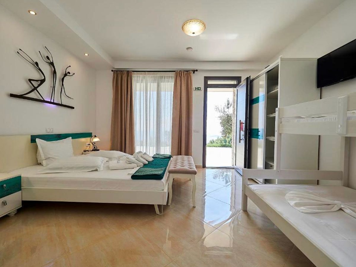 A bedroom with a white double bed and teal headboard, a cushioned bench at its foot and a set of white bunk beds, with a glass door open onto the garden.