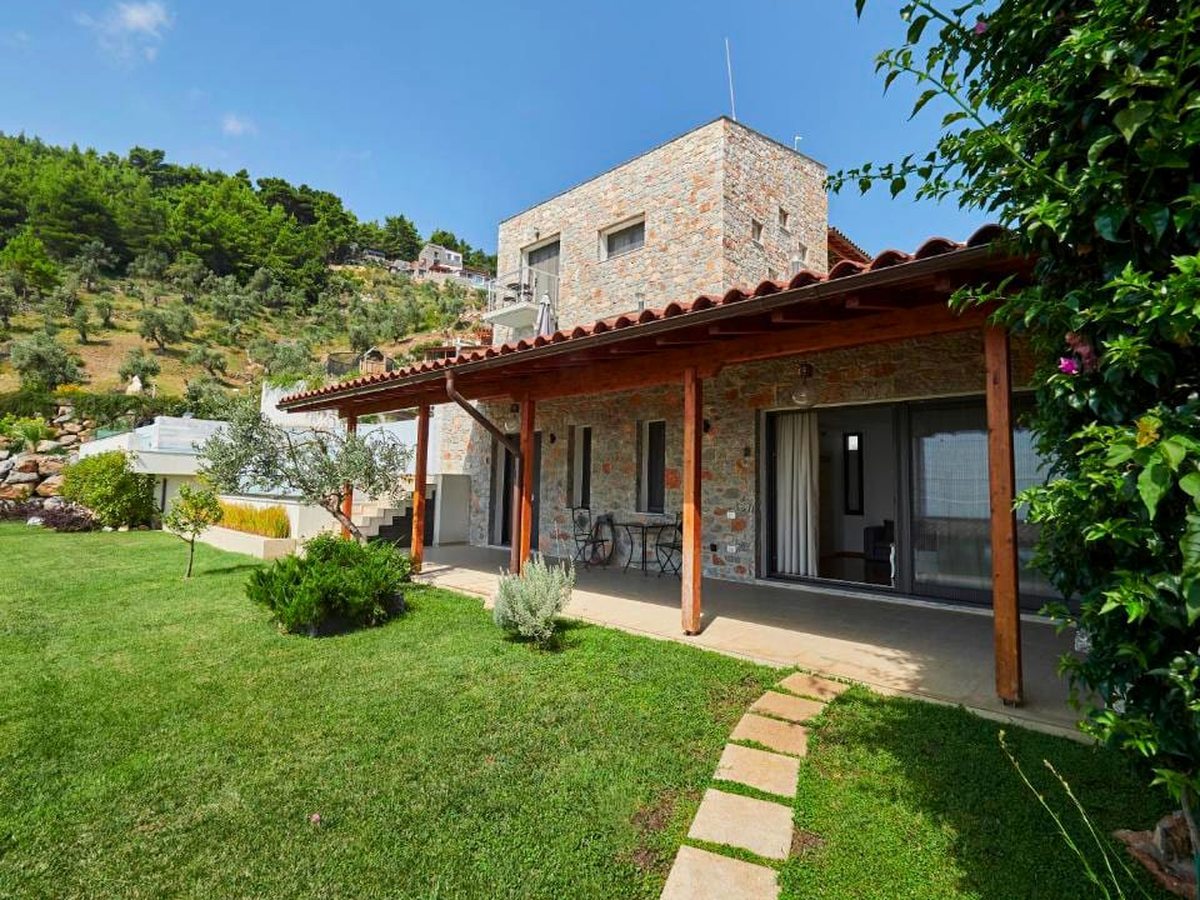 A single-storey stone villa with a red-tiled roof and a covered wooden veranda, standing on a mown lawn with olive trees and a pine-covered hillside behind.