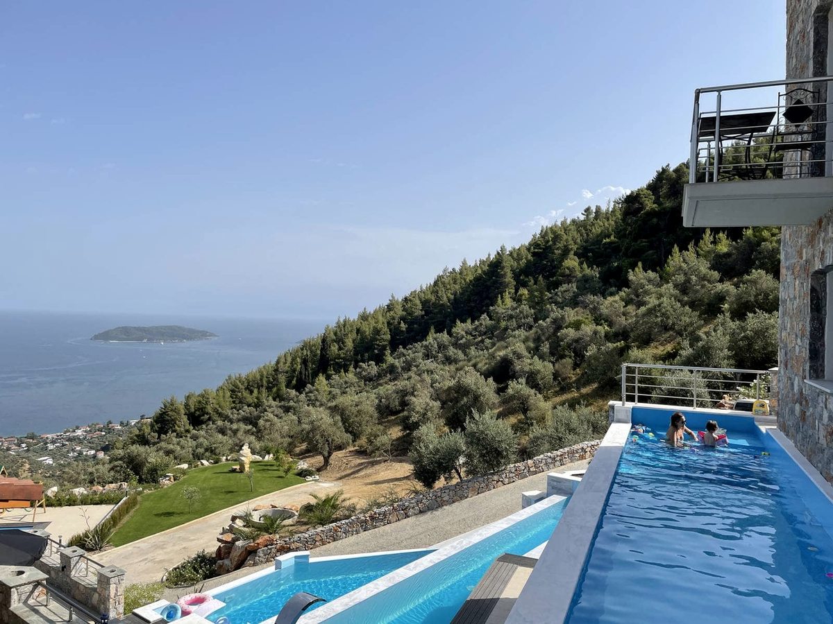 Two blue infinity pools on stepped terraces beside a stone villa, with a woman and a child in the upper pool and a wooded hillside dropping to the sea.