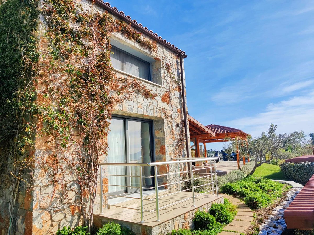 The side of a stone villa covered in climbing plants, with sliding glass doors opening onto a small decked terrace behind a steel railing.