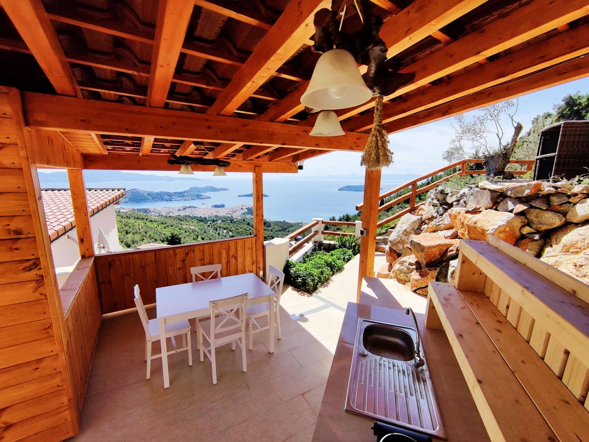 A covered timber terrace with a stainless-steel outdoor kitchen unit and sink, a white dining table and chairs and a long bench, looking out over pine hills to the sea.