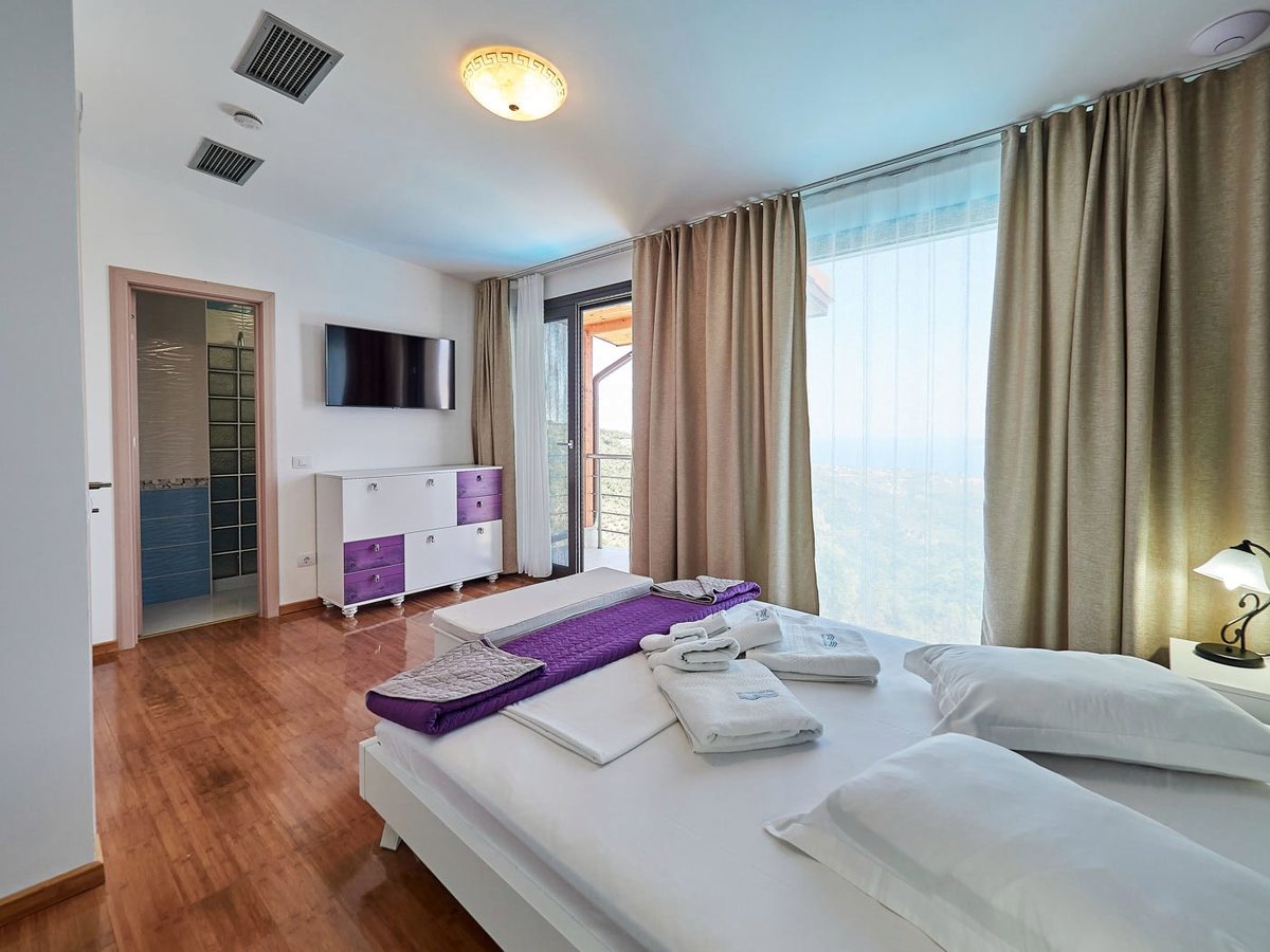 A bedroom with a wide white bed, a chest of drawers, a wall-mounted television and an open door through to a tiled en-suite bathroom, with balcony doors to one side.