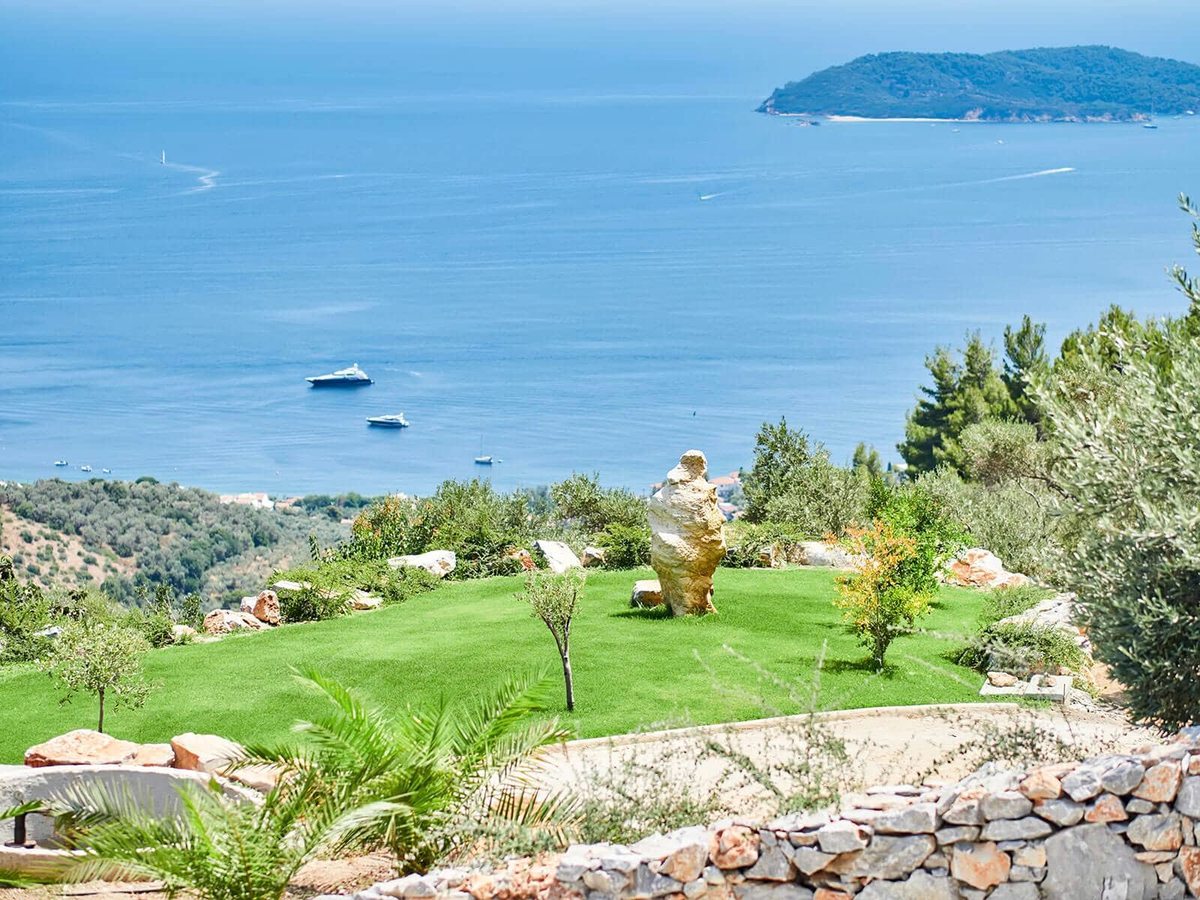 A mown lawn edged with young olive trees and a carved stone sculpture, dropping away to blue sea and a wooded islet.