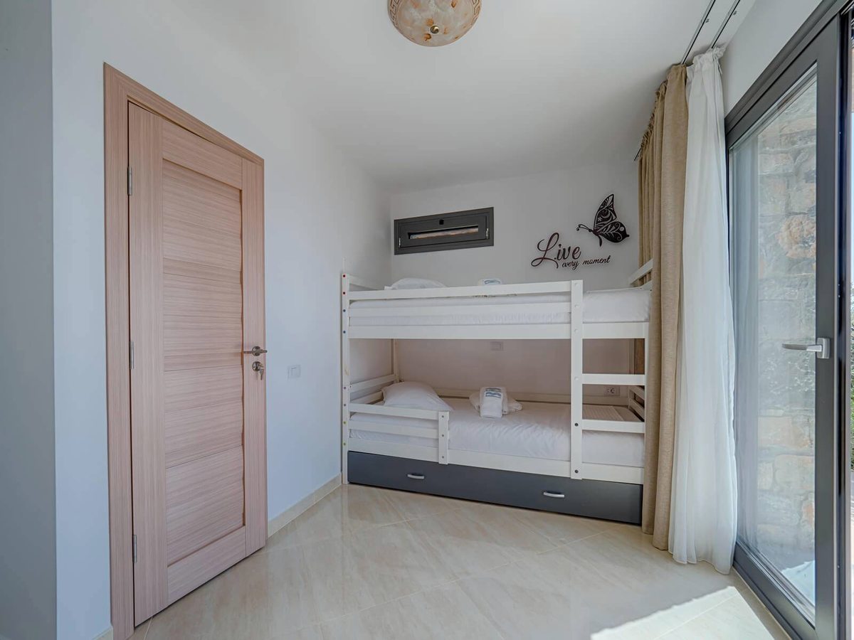 A small bedroom with white bunk beds over a pull-out drawer bed, a pale wooden door and a sliding glass door to a stone-walled terrace.