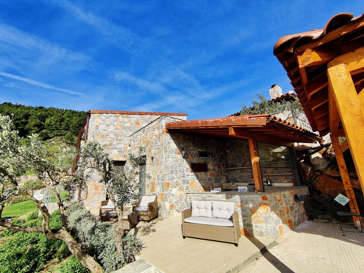 A stone-built villa courtyard with wicker sofas and armchairs on a paved terrace under a timber pergola, with olive trees to one side.