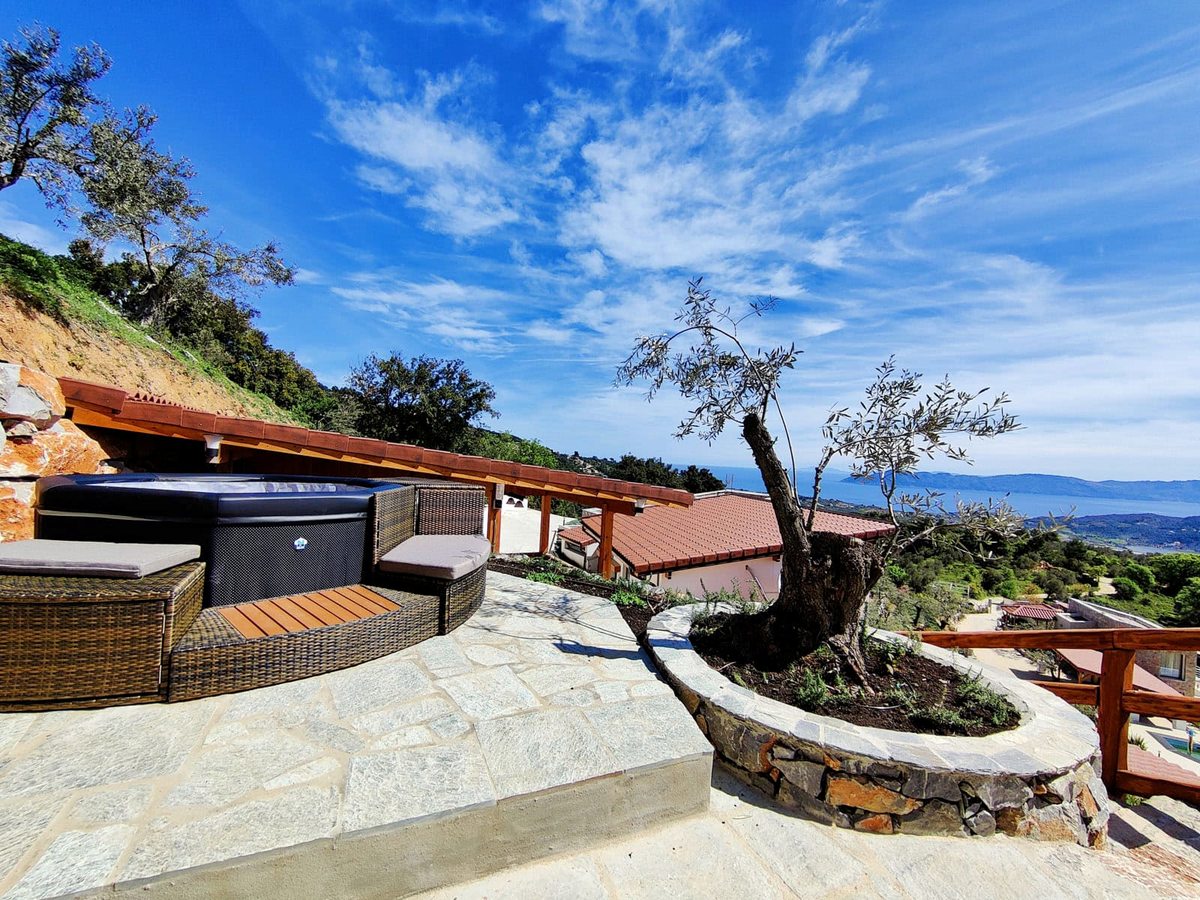 An outdoor hot tub in a rattan surround with its cover closed and wicker seating around it on a stone-paved terrace, framed by an old olive tree, with the sea and rooftops beyond.