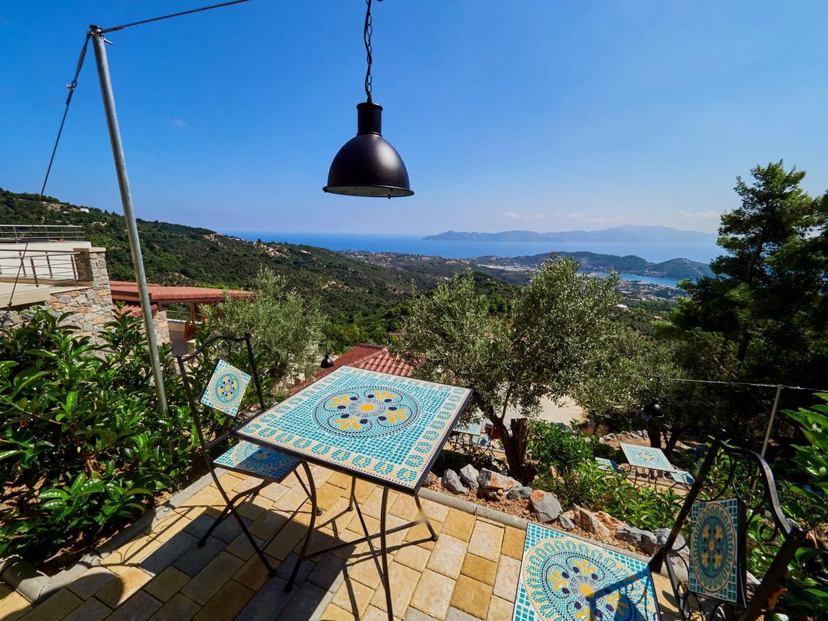 Folding metal chairs and tables with blue and yellow mosaic tops on a paved terrace under a black pendant lamp, with olive trees and the sea beyond.