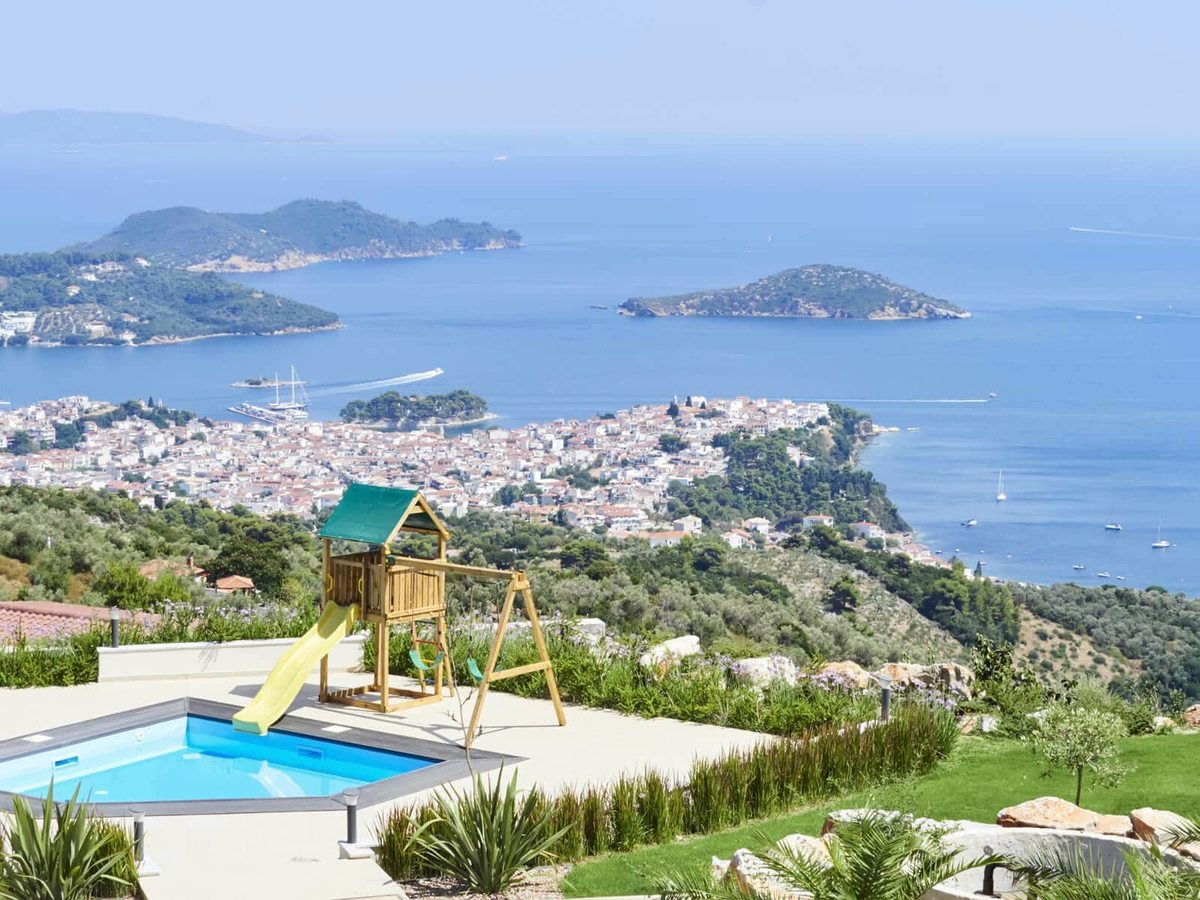 A small square children's pool beside a wooden play frame with a yellow slide and two swings, on a paved terrace high above Skiathos Town and the sea.