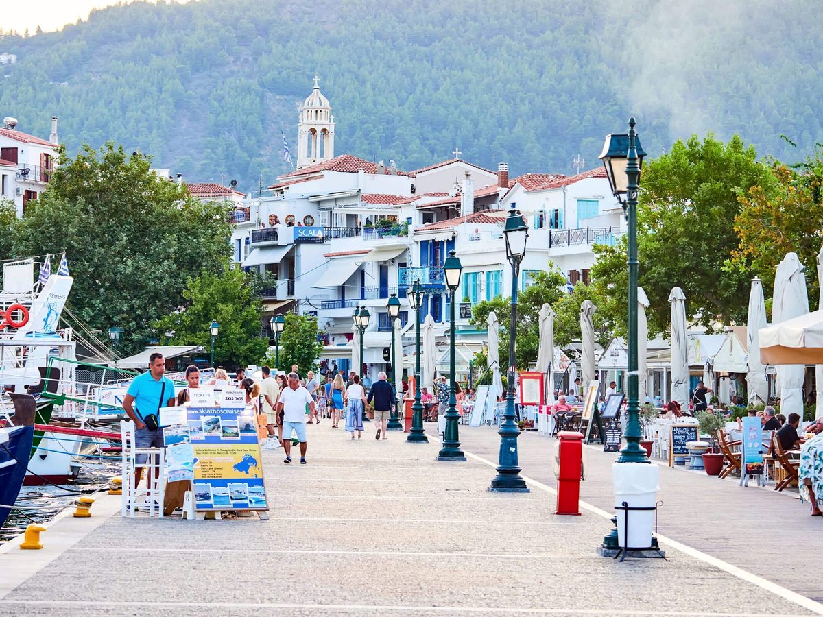 A wide paved harbour promenade with green lamp posts, cafe tables and moored boats, and white houses with terracotta roofs rising to a church tower and wooded hill.