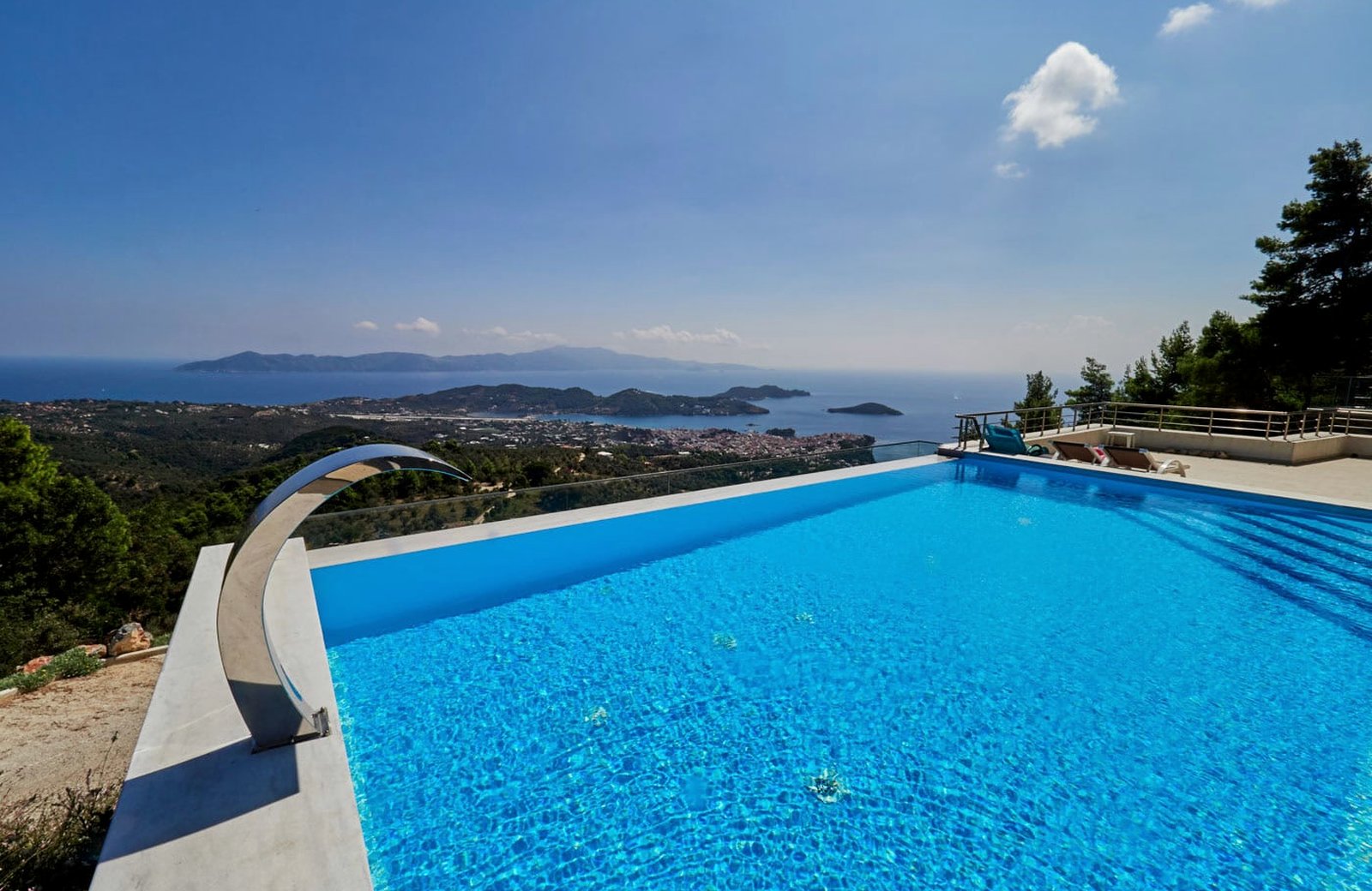 An infinity pool at Sky Sea Resort & Villas, its water running level with the terrace edge, above wooded hills with Skiathos Town, its harbour and the islets beyond.