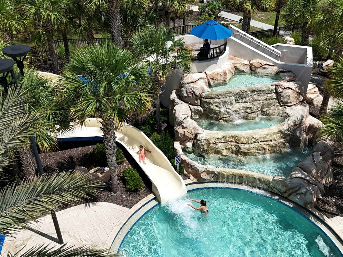 A white open waterslide curving down a tall faux-rock waterfall structure into a round pool, with a rider partway down and palms overhead.