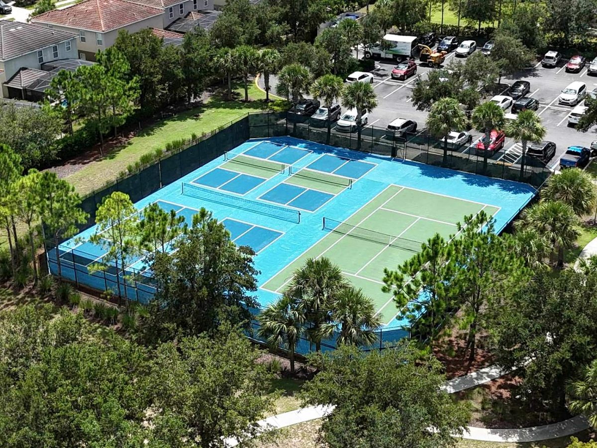 An aerial view of blue and green hard courts marked for tennis and pickleball, surrounded by mature trees, footpaths, parked cars and resort villas.