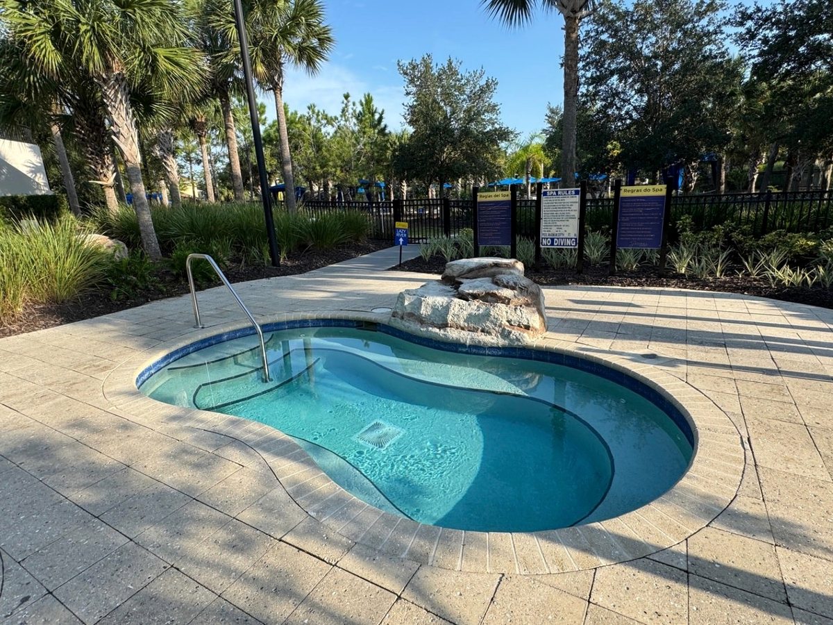 A kidney-shaped outdoor hot tub set into a paved deck with a rock water feature, edged by a safety fence and tropical planting.