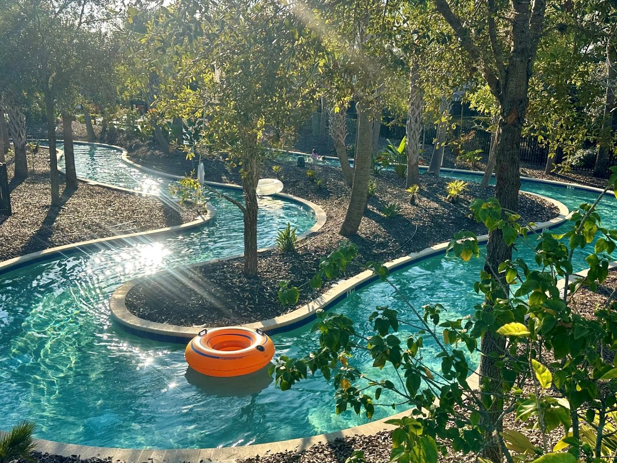 A winding blue lazy river channel with an orange inflatable ring floating on it, threading between planted islands and mature trees in dappled sunlight.