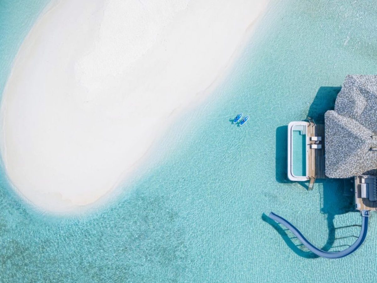 Aerial view of an overwater villa with a private pool and a curved slide dropping into a shallow turquoise lagoon beside a white sandbank.