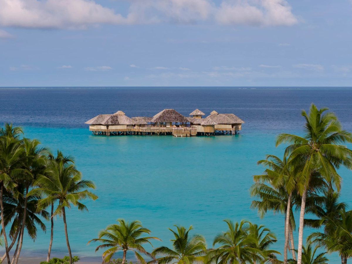 A cluster of thatched-roof buildings standing on stilts above a turquoise lagoon, framed by palm trees under a blue sky.