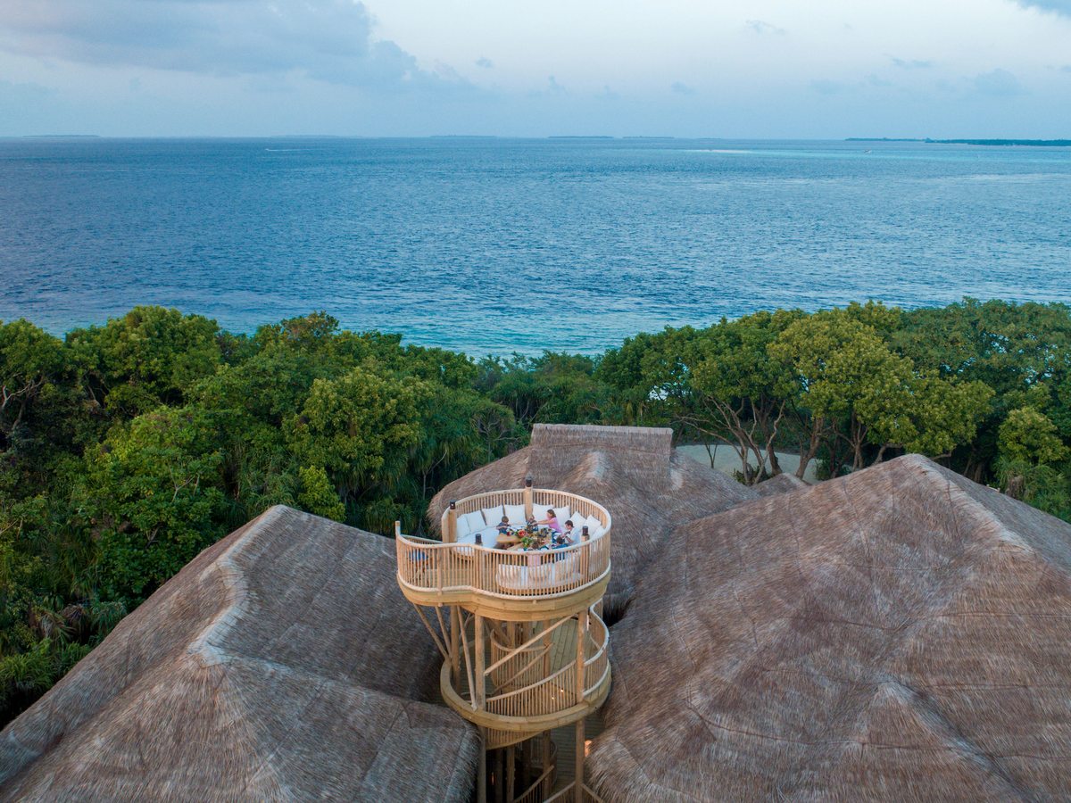 A small circular dining deck on a wooden tower rising above thatched villa roofs, with jungle and open sea beyond.