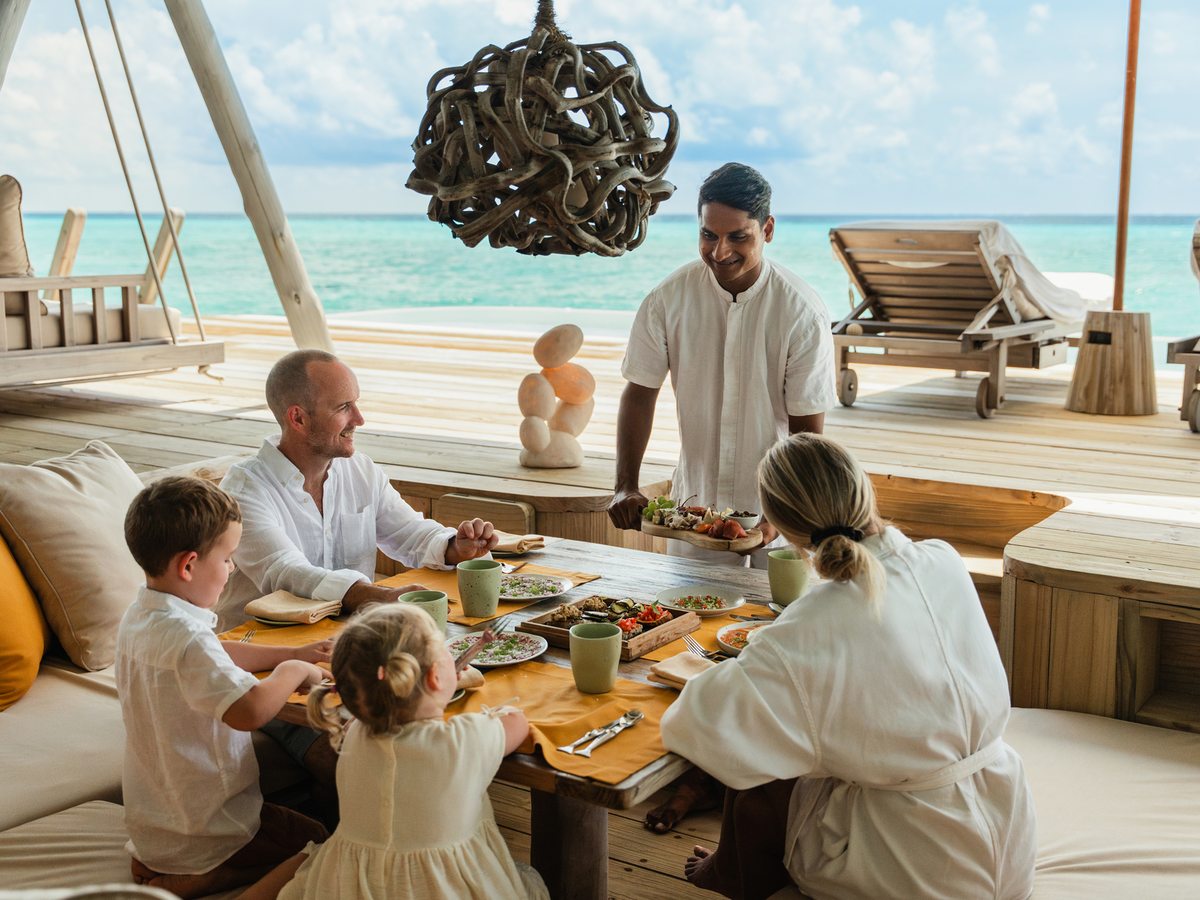 A man, a woman and two young children eating at a low wooden table on an open villa deck while a host in white serves a platter, with the lagoon behind.