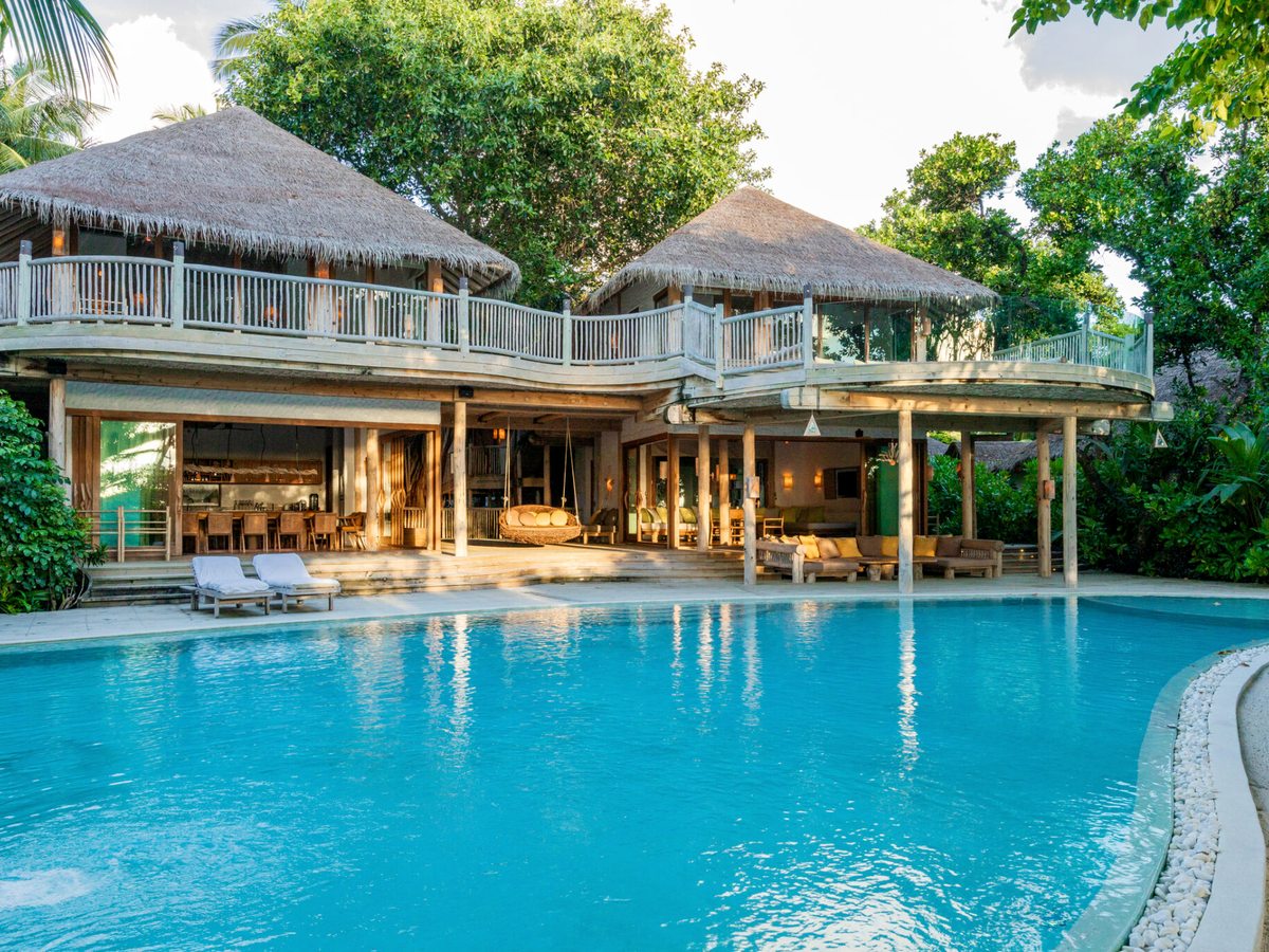 A large two-storey thatched villa with open living areas at ground level and a wide curving blue pool in front.