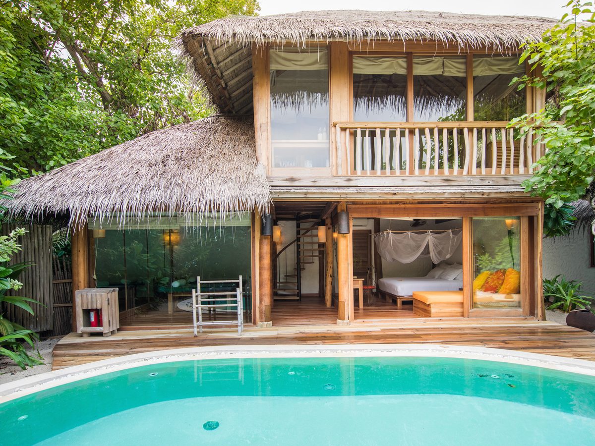 A two-storey thatched villa with open glass walls, a canopy bed on the ground floor and a curved pool in front, surrounded by trees.