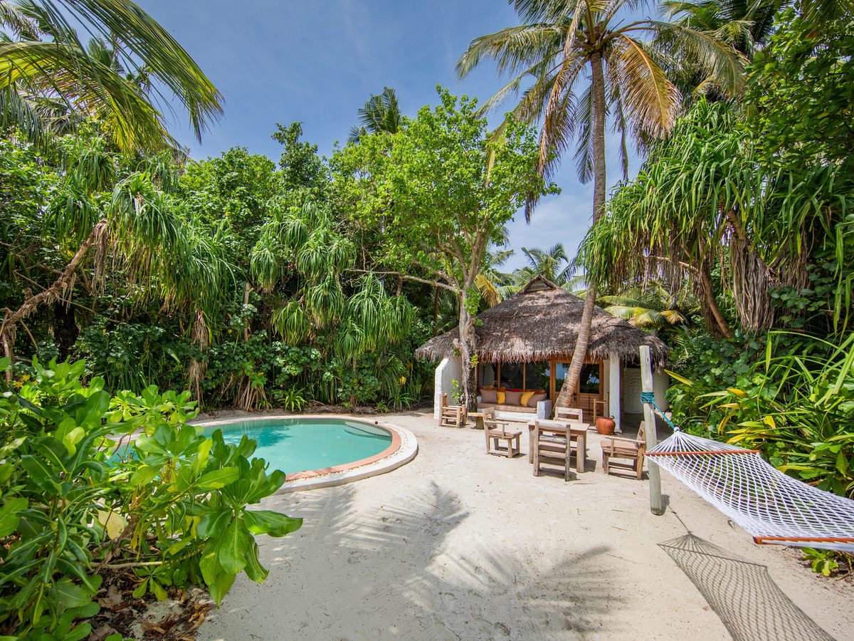 A small round pool beside a sandy courtyard with a thatched cabana, wooden seats and a hammock among palms.