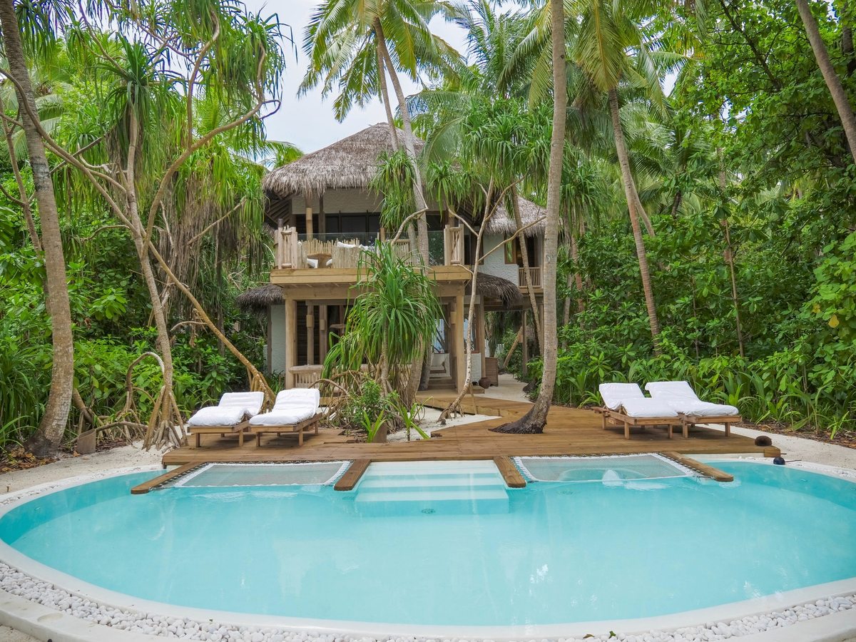 A large round pool with a wooden deck and sun loungers in front of a two-storey thatched villa surrounded by tall palms.