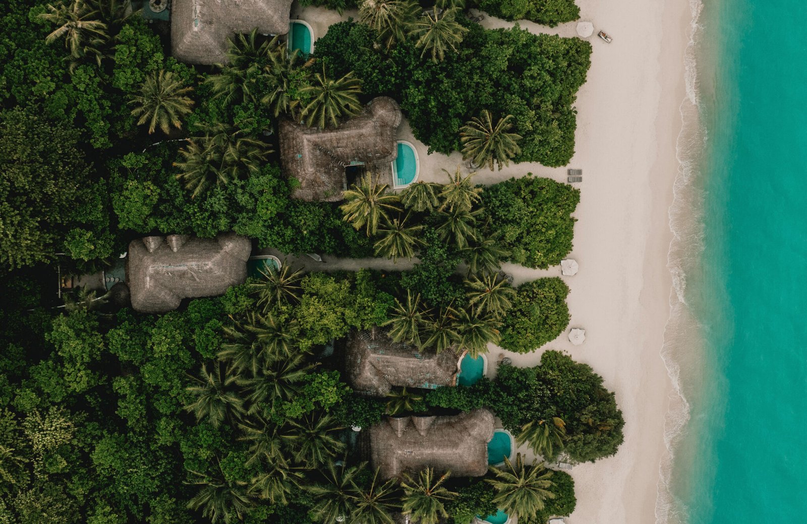 Aerial view of thatched Soneva Fushi villas with private pools set in dense jungle along a wide white beach, with the turquoise Baa Atoll lagoon beyond.