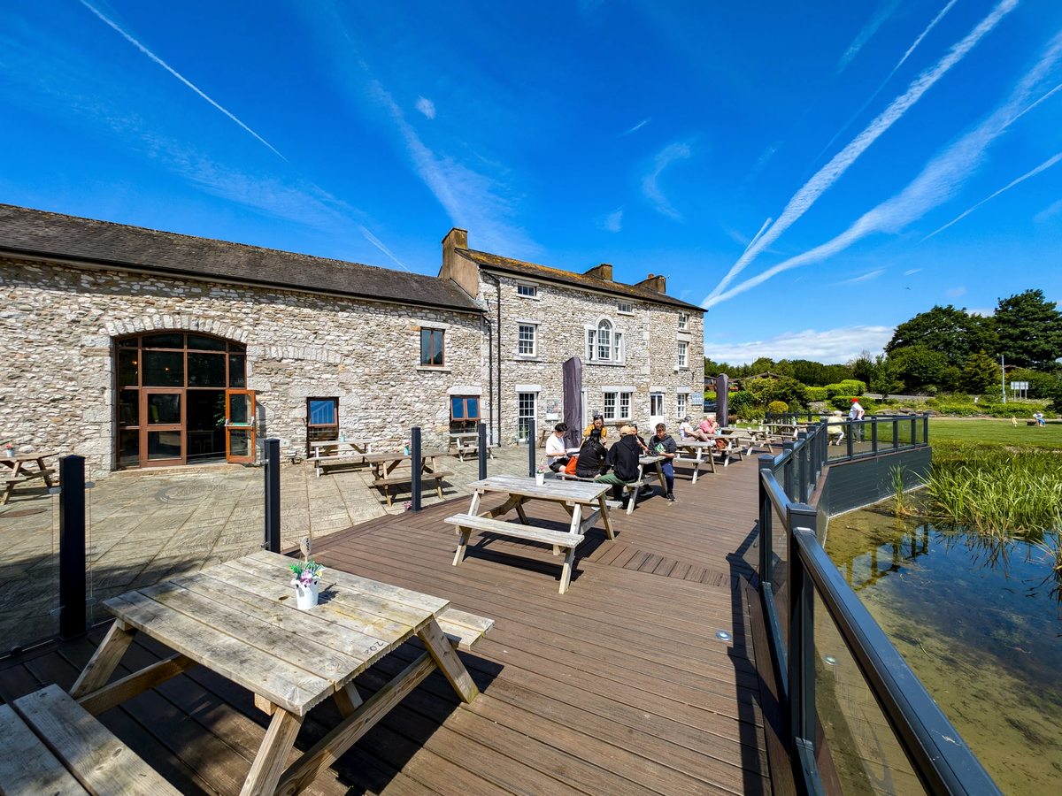 A wide timber deck of picnic benches beside the water with people eating and drinking, in front of a long stone building under a blue sky.