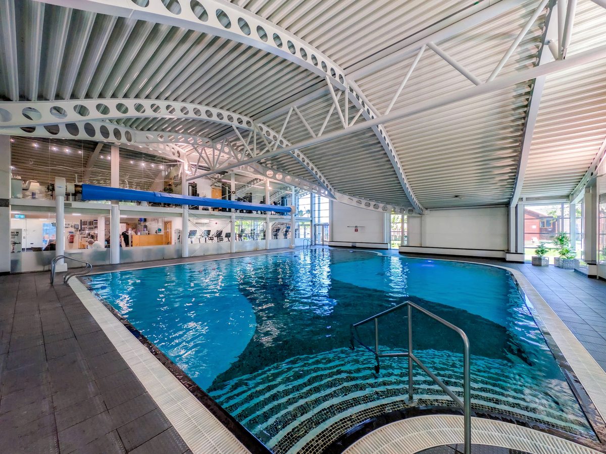 An indoor swimming pool with blue water under a curved arched roof, with a tiled surround and steps and handrails at the near end.