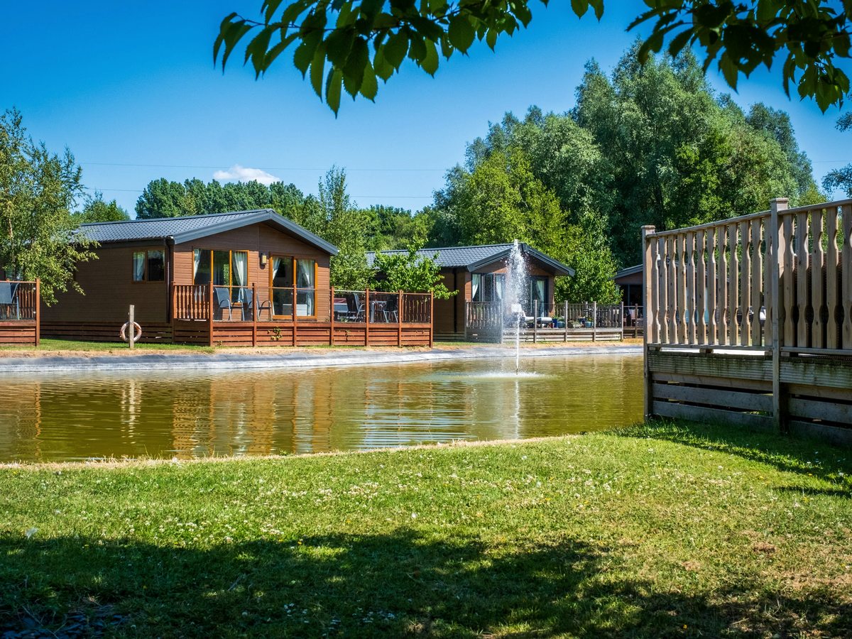 Two timber lodges with balustraded verandas and patio furniture beside a small stretch of water with a fountain, backed by trees.