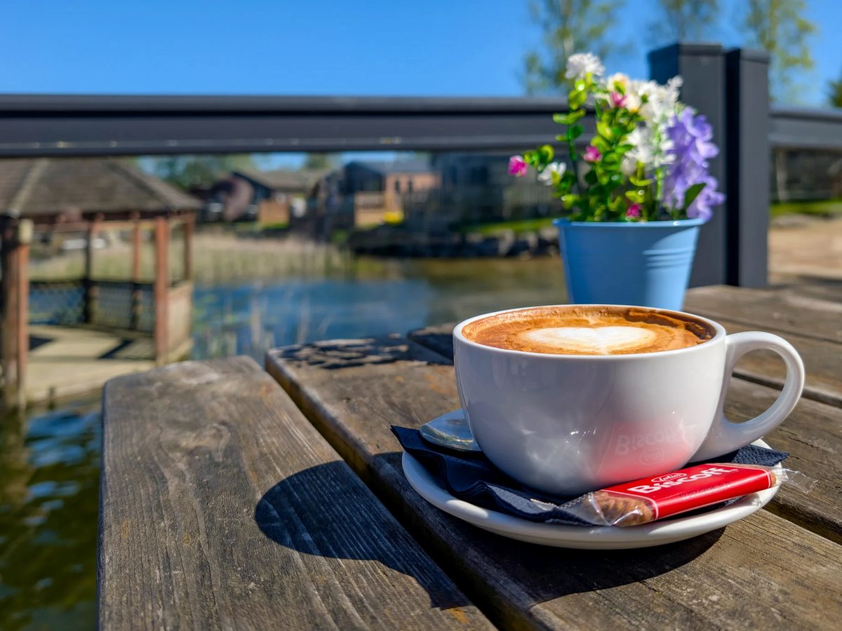 A cappuccino in a white cup and saucer on a wooden table on the deck, with a pot of flowers beside it and the water and lodges beyond.